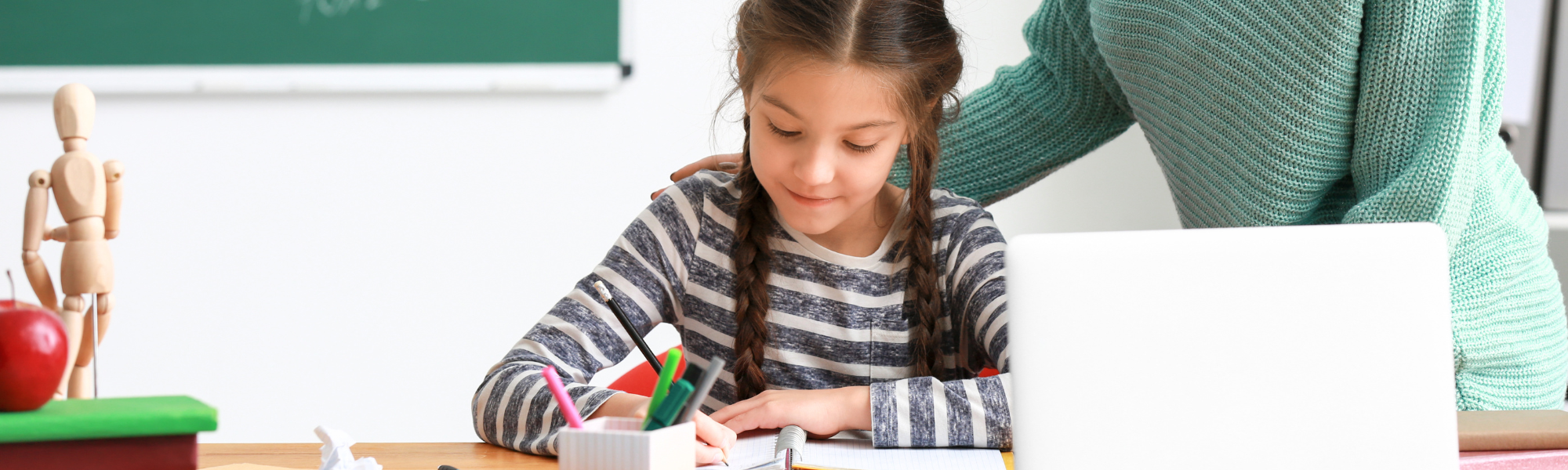 Meisje schrijft ontspannen in haar schrift terwijl de leerkracht achter haar staat met een hand op haar schouder