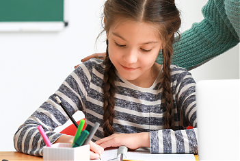 Meisje schrijft ontspannen in haar schrift terwijl de leerkracht achter haar staat met een hand op haar schouder