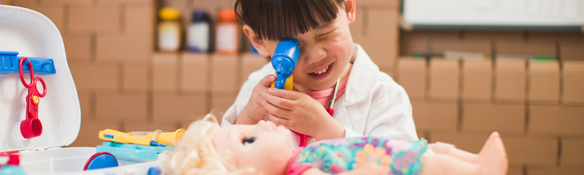 Een kleuter in doktersjas met pop en dokterskoffer doet onderzoek