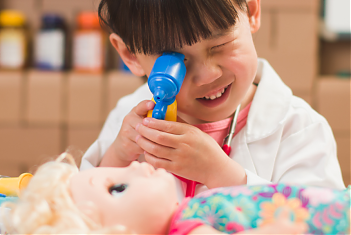 Een kleuter in doktersjas met pop en dokterskoffer doet onderzoek
